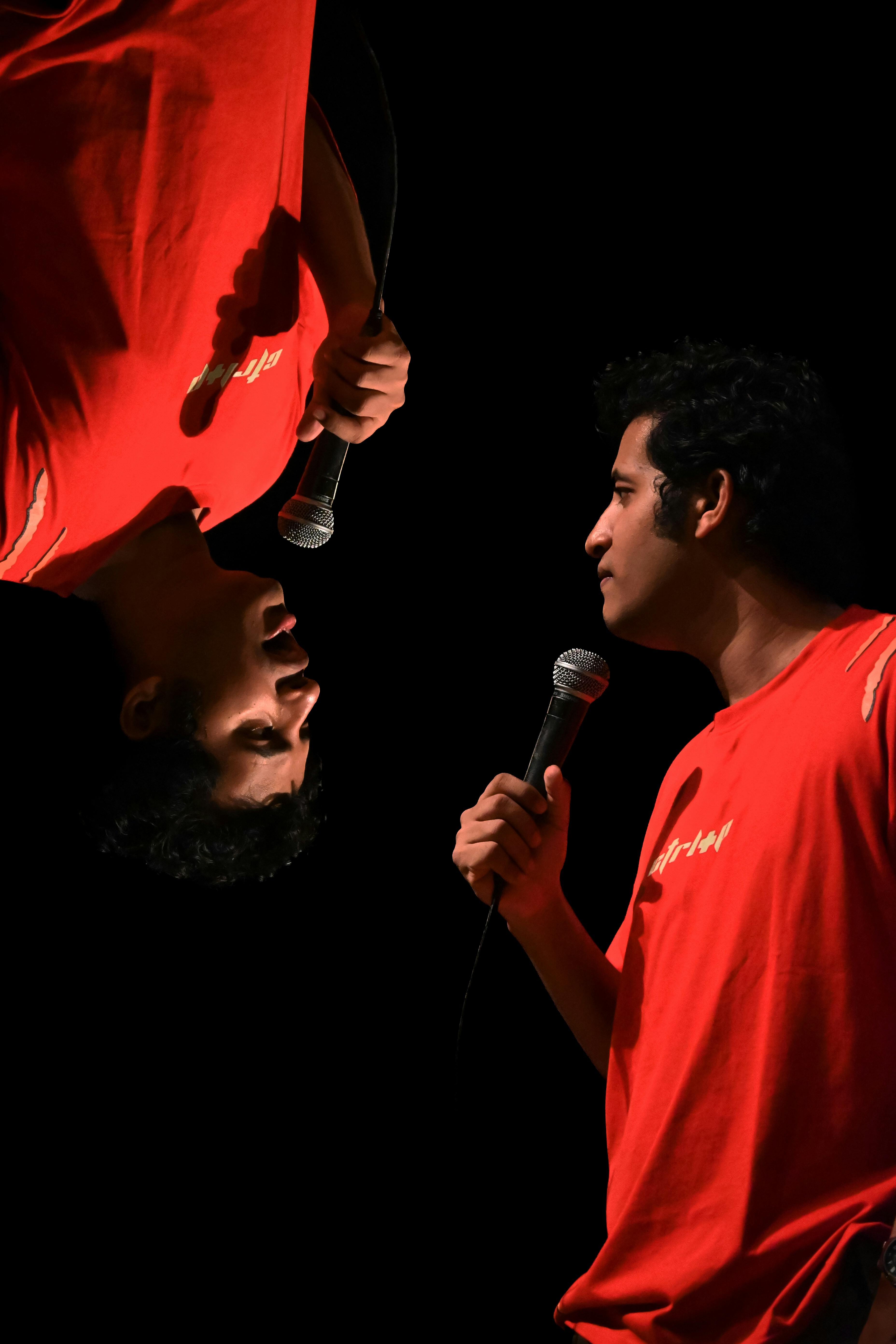 A performer stands alone, doubled in quiet thought. by School Play Scripts Man in red shirt holding microphone, upside down reflection.