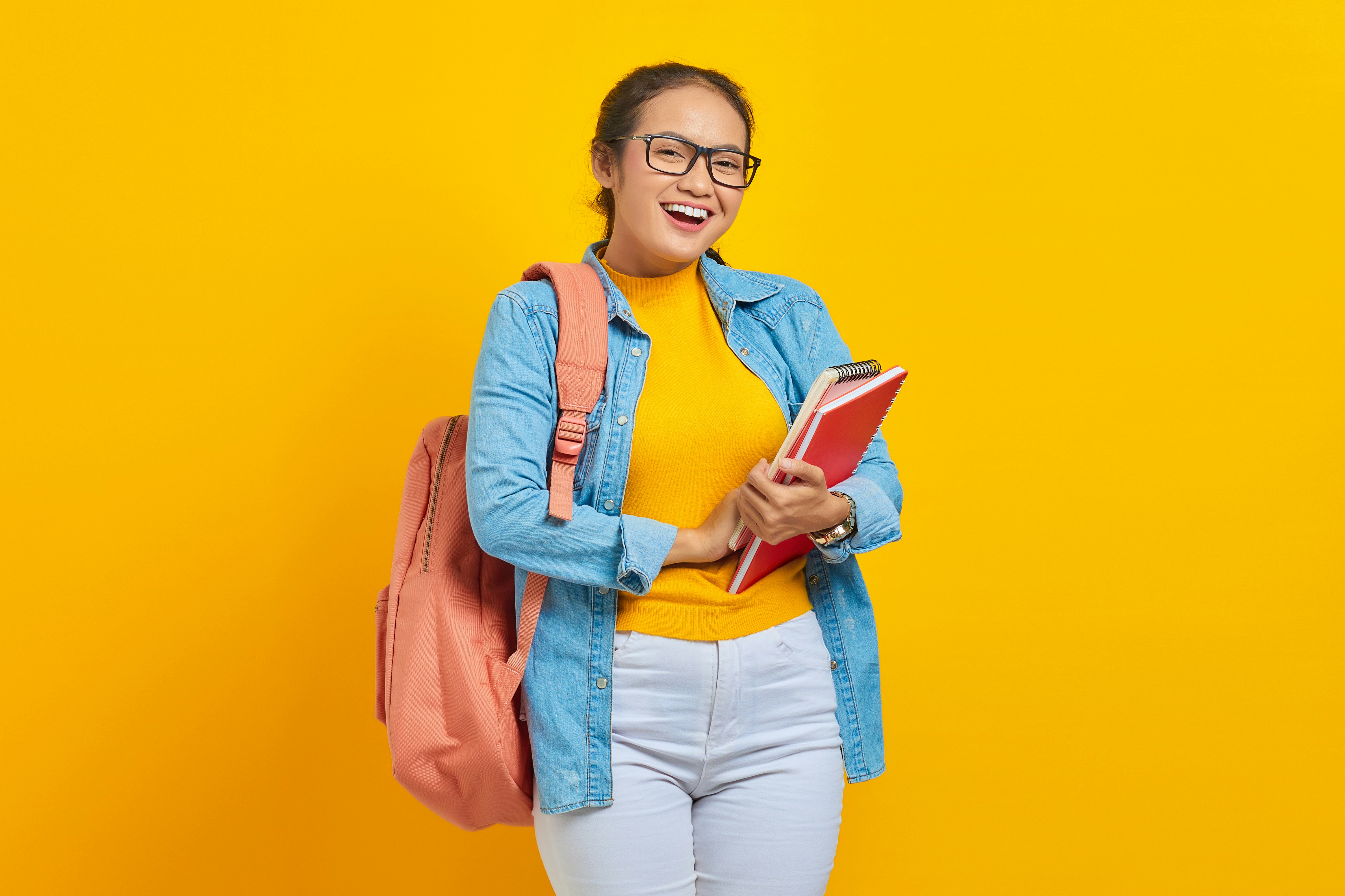 Portrait of cheerful young Asian woman student in casual clothes with backpack holding book and looking at camera isolated on yellow background by School Play Scripts a woman with a backpack and a book smiling